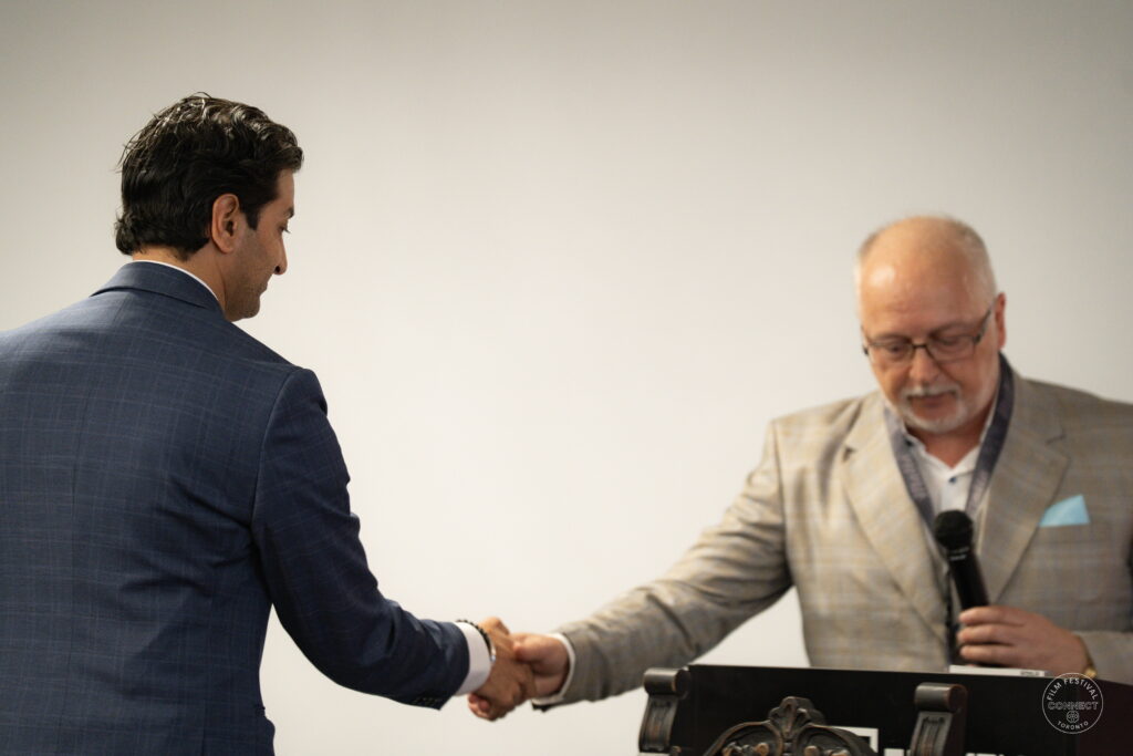 Two speakers shake hands on stage, with one holding a microphone at the AI & Film Forum.