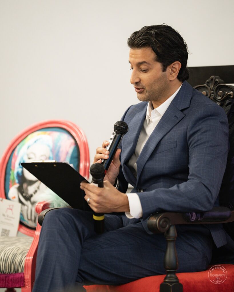The moderator speaks into a microphone while consulting notes on his lap. AI & Film Forum, Panel 1, September 14, 2026.