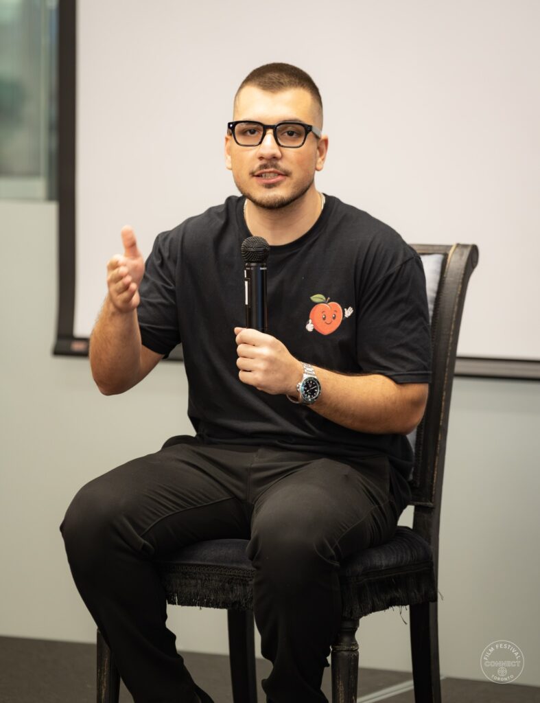 A panel participant wearing glasses and a black T-shirt holds a microphone and gestures toward the audience. AI & Film Forum, Panel 3, September 14, 2026.