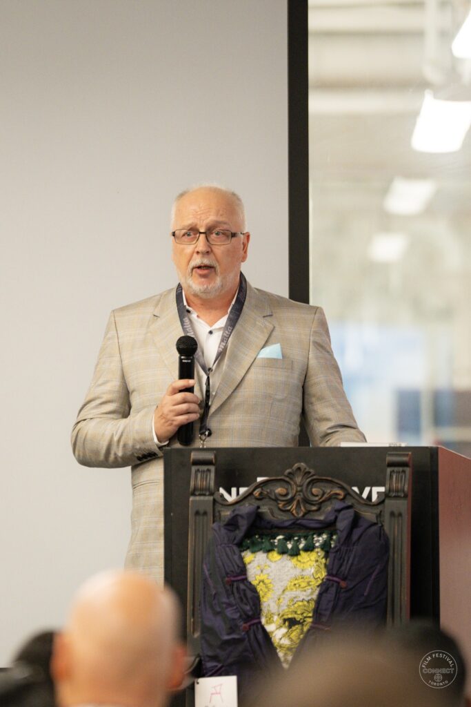 A speaker with glasses addresses the room with a microphone behind a decorative chair.