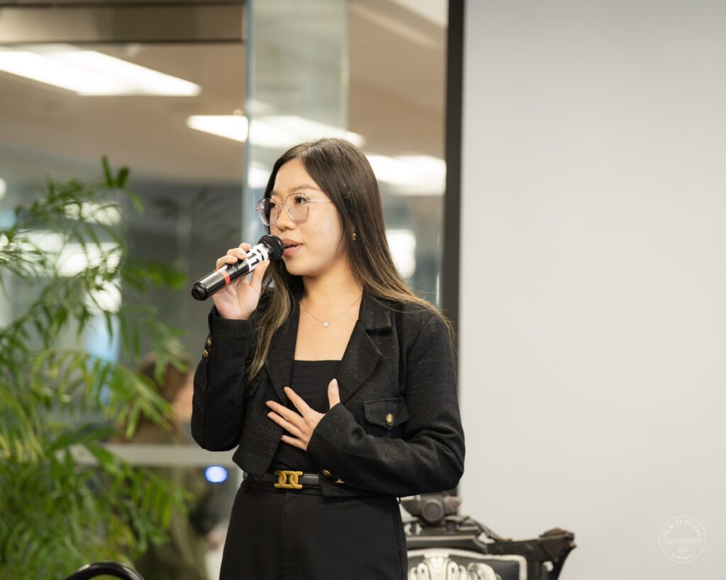 A woman in a black jacket speaks into a microphone and gestures near the stage.