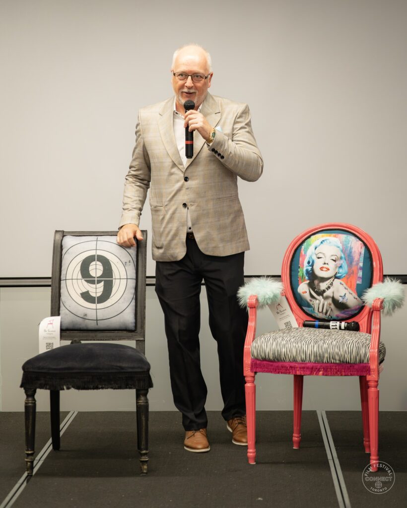 A speaker in a light jacket holds a microphone between decorative stage chairs.